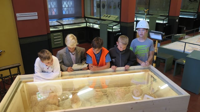 Fünf Jungs schauen in eine Glasvitrine, in welcher sich ein 2000 Jahre alter Grabstein befindet. Im Hintergrund ist das Museum zu sehen. Fünf Jungs schauen in eine Glasvitrine, in welcher sich ein 2000 Jahre alter Grabstein befindet. Im Hintergrund ist das Museum zu sehen.