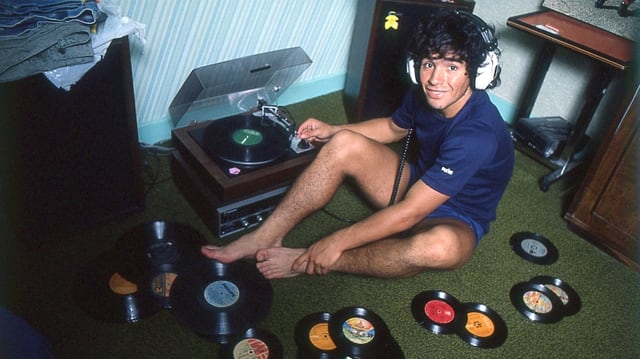 Ein junger Maradona sitzt auf dem Fussboden neben einem Plattenspieler. Um ihn herum sind viele Vinyl-Platten, er hat Kopfhörer auf. Ein junger Maradona sitzt auf dem Fussboden neben einem Plattenspieler. Um ihn herum sind viele Vinyl-Platten, er hat Kopfhörer auf.