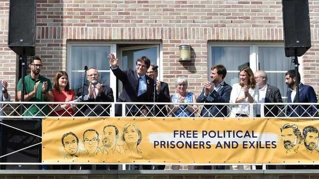 Leute auf einer Terrasse. Puigdemont grüsst mit erhobener Hand. Ein Plakat fordert die Freilassung aller politischer Gefangener und Exilanten. Leute auf einer Terrasse. Puigdemont grüsst mit erhobener Hand. Ein Plakat fordert die Freilassung aller politischer Gefangener und Exilanten.
