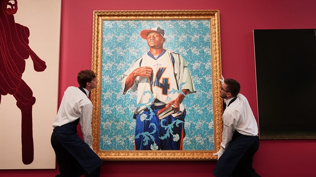 Ein gerahmtes Bild zeigt einen jungen schwarzen Mann in Hip-Hop-Kleidung und mit einem Buch in der Hand. Zwei Mitarbeiter eines Museums hängen es auf. Ein gerahmtes Bild zeigt einen jungen schwarzen Mann in Hip-Hop-Kleidung und mit einem Buch in der Hand. Zwei Mitarbeiter eines Museums hängen es auf.