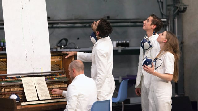 Vier Menschen in weissen Anzügen: Drei stehen und blicken in die Höhe, einer sitzt am Klavier. Vier Menschen in weissen Anzügen: Drei stehen und blicken in die Höhe, einer sitzt am Klavier.