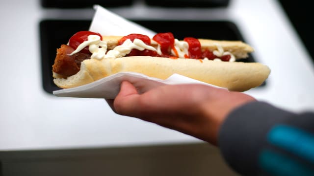 Eine Hand hält einen geschlitzten Hotdog, aus dem das Ketchup quillt. Eine Hand hält einen geschlitzten Hotdog, aus dem das Ketchup quillt.