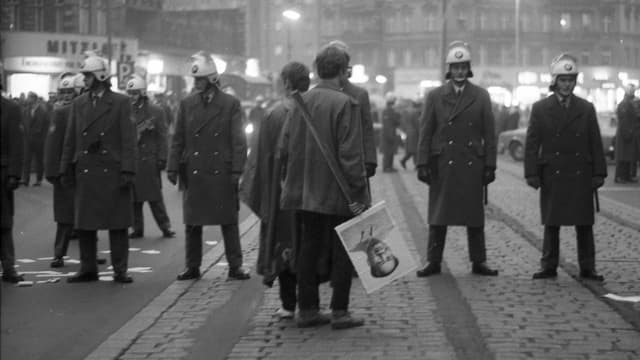 Schwarzweiss-Bild: ein Demonstrant mit einem Mao-Plakat vor einer Reihe behelmter Polizisten. Schwarzweiss-Bild: ein Demonstrant mit einem Mao-Plakat vor einer Reihe behelmter Polizisten.