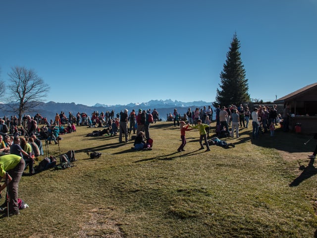 Auf einer Wiese sitzen Personen auf Bänken oder auf dem Grass und spielen, essen oder bestaunen einfach die herrliche Aussicht auf die Bergwelt.