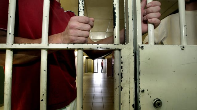 Symbolbild eines Mannes, der hinger Gitterstäben im Gang eines Gefängnisses steht. Zu sehen sind die Hände, die Arme und der Oberkörper. Symbolbild eines Mannes, der hinger Gitterstäben im Gang eines Gefängnisses steht. Zu sehen sind die Hände, die Arme und der Oberkörper.