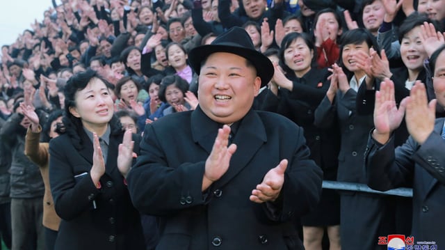 Kim in schwarzem Mantel mit Hut klatscht und lacht vor einer begeisterten Menschenmenge. Kim in schwarzem Mantel mit Hut klatscht und lacht vor einer begeisterten Menschenmenge.