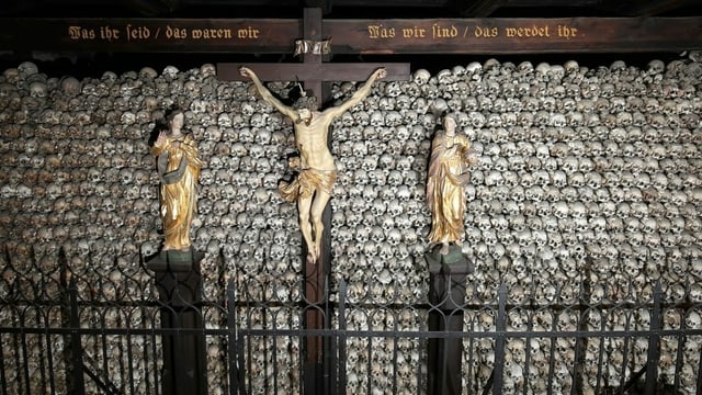 Unzählige, aufeinandergsetapelte Totenschädel. Davor Christuskreuz, darüber ein Holzbalken mit Inschrift. Unzählige, aufeinandergsetapelte Totenschädel. Davor Christuskreuz, darüber ein Holzbalken mit Inschrift.