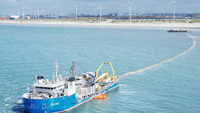 Schiff zieht ein zu verlegendes Hochseekabel hinter sich her, im Hintergrund eine Küste mit Windrädern zur Stromproduktion Schiff zieht ein zu verlegendes Hochseekabel hinter sich her, im Hintergrund eine Küste mit Windrädern zur Stromproduktion