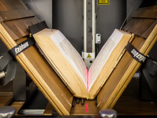 Buch liegt halb geöffent in einer Maschine; rote Linie eines Lasers ist im Falz sichtbar. Buch liegt halb geöffent in einer Maschine; rote Linie eines Lasers ist im Falz sichtbar.