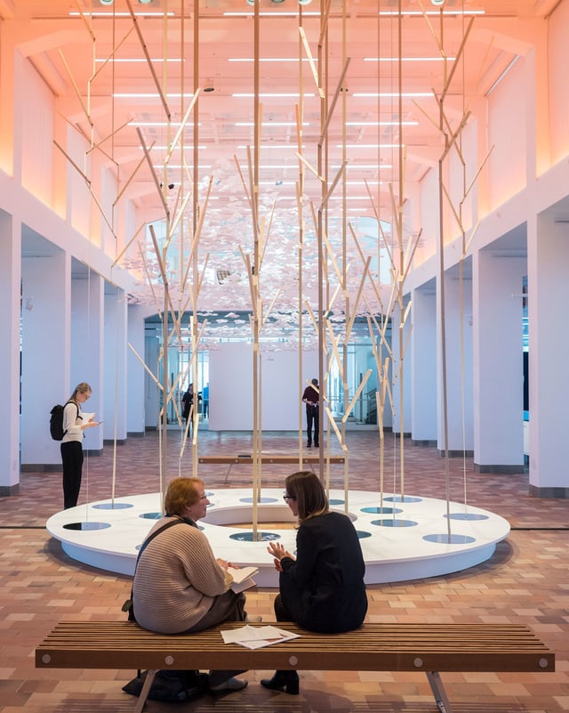 Zwei Museumsbesucherinnen sitzen vor einer Skulptur aus Holzstäben. Zwei Museumsbesucherinnen sitzen vor einer Skulptur aus Holzstäben.
