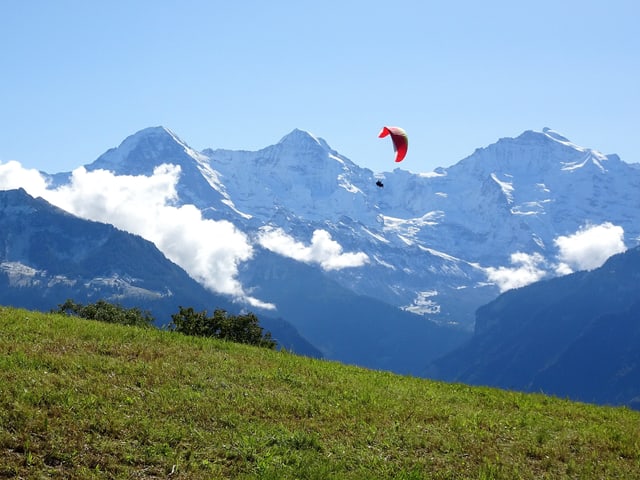 Ein Gleitschirmflieger gleitet hoch oben am blauen Himmel. Die Berge sind frisch verschneitl.