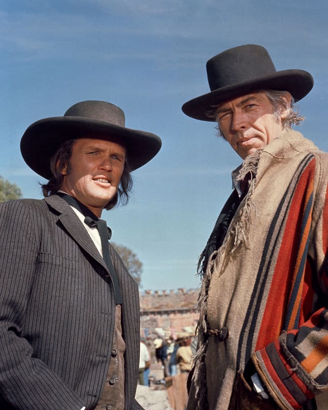 Filmstill aus einem Western: zwei lächelnde Männer mit schwarzem Hut.