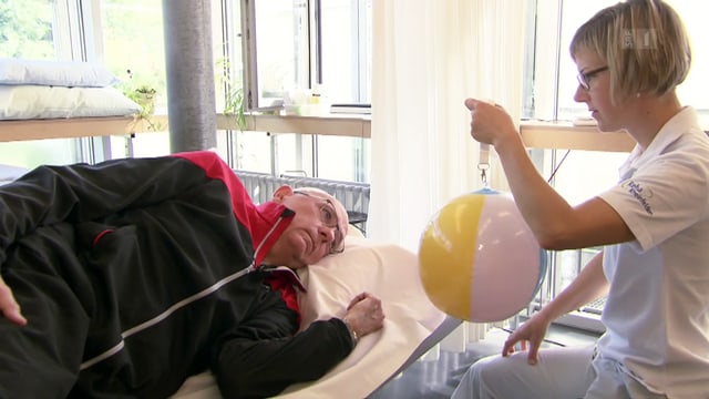 Ein Patient liegt auf einer Liege, die Therapeutin sitzt vor ihm mit einem bunten Ball. Ein Patient liegt auf einer Liege, die Therapeutin sitzt vor ihm mit einem bunten Ball.