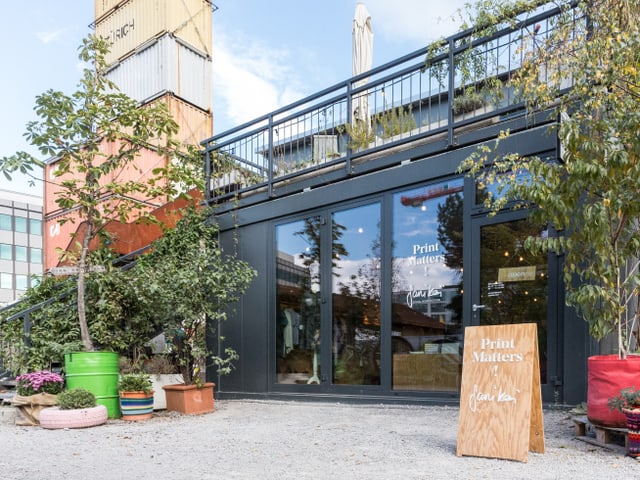 Glasfront eines Ladenprovisoriums, im Hintergrund ein Turm aus Schiffscontainern. Glasfront eines Ladenprovisoriums, im Hintergrund ein Turm aus Schiffscontainern.
