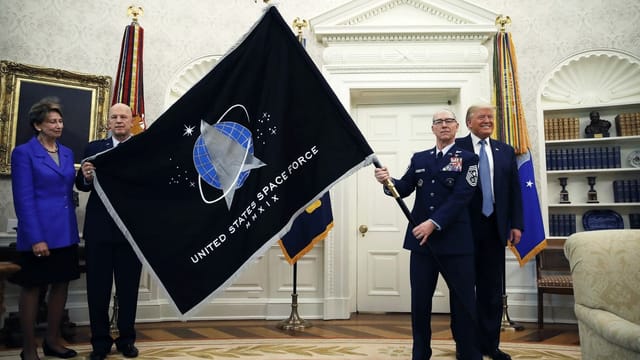 Das Oval Office im Weissen Haus. Ein Mann in Unoform schwenk eine riesige Fahne. Sie ist schwarz, darauf ein blauer Kreis und davor ein silbernes Dreieck. Im Hintergrund lacht Donald Trump. Das Oval Office im Weissen Haus. Ein Mann in Unoform schwenk eine riesige Fahne. Sie ist schwarz, darauf ein blauer Kreis und davor ein silbernes Dreieck. Im Hintergrund lacht Donald Trump.