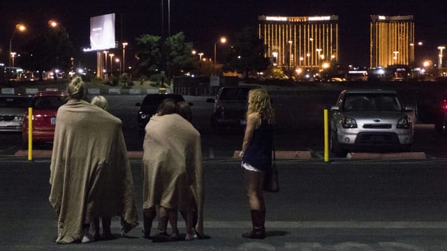 Mehrere Personen stehen mit dem Rücken zur Kamera, einige haben Decken über den Schultern, in der Ferne das beleuchtete Hotel Mandalay Bay. Mehrere Personen stehen mit dem Rücken zur Kamera, einige haben Decken über den Schultern, in der Ferne das beleuchtete Hotel Mandalay Bay.