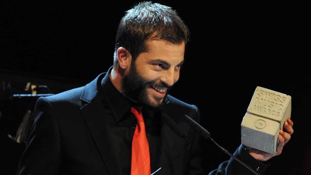 Zu sehen ist der Musiker Bligg. Er trägt schwarzen Anzug, schwarzes Hemd, schwarzen Bart und er lächelt. In der linken Hand hält er den Betonklotz, der den Swiss Music Award darstellt. Zu sehen ist der Musiker Bligg. Er trägt schwarzen Anzug, schwarzes Hemd, schwarzen Bart und er lächelt. In der linken Hand hält er den Betonklotz, der den Swiss Music Award darstellt.