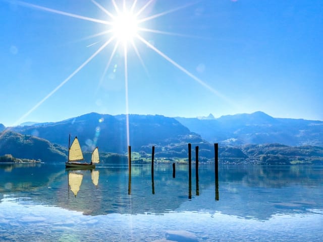 Die Sonne steht am wolkenlosen Himmel. Vor einer Hügelkette liegt ein See mit glatter Oberfläche. Auf dem See ist ein Segelschiff.