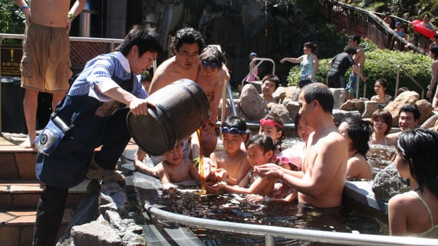 In einem Bassin sitzen eine Gruppe Kinder und Erwachsene und halten ihre Hände aus. Ein Angestellter leert ihnen Kaffee ins Badewasser. In einem Bassin sitzen eine Gruppe Kinder und Erwachsene und halten ihre Hände aus. Ein Angestellter leert ihnen Kaffee ins Badewasser.