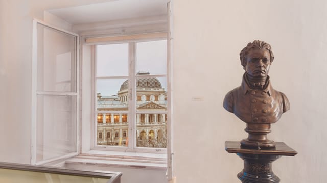 Im Vordergrund steht eine Statue. Im Hintergrund sieht man eine Art Palast durch ein Fenster. Im Vordergrund steht eine Statue. Im Hintergrund sieht man eine Art Palast durch ein Fenster.
