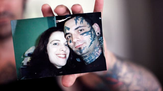 Verschwommen im Hintergrund sieht man einen Mann. Er hält ein Foto in die Kamera. Darauf zu sehen ist er selbst mit Tattoos im Gesicht. Neben ihm eine lächelnde Frau.