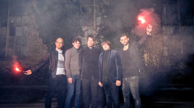 Die fünf Band-Mitglieder stehen aufgereiht vor einem Gebäude, zwei der Männer haben eine brennende Fackel in der Hand. Die fünf Band-Mitglieder stehen aufgereiht vor einem Gebäude, zwei der Männer haben eine brennende Fackel in der Hand.