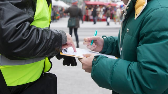 Ein Herr sammelt auf der Strasse Unterschriften, eine Frau unterschreibt einen Papierbogen. Ein Herr sammelt auf der Strasse Unterschriften, eine Frau unterschreibt einen Papierbogen.