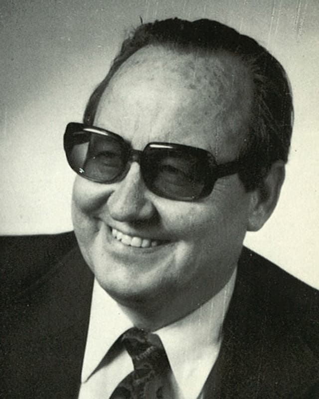 Schwarz-weisse Fotografie mit Porträt von Jost Marty mit Anzug und Krawatte und grosser dunkler Brille.