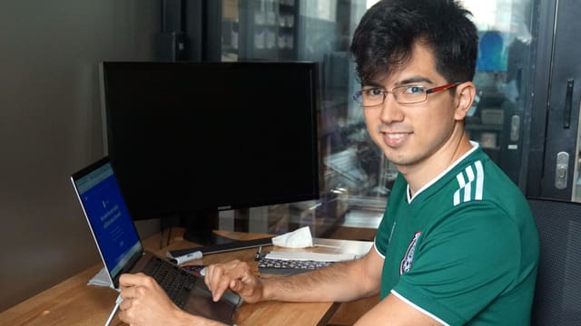 Ein junger Mann mit einem Fussballtrikot, dunklen Haaren und einer Brille sitzt vor einem Laptop. Ein junger Mann mit einem Fussballtrikot, dunklen Haaren und einer Brille sitzt vor einem Laptop.