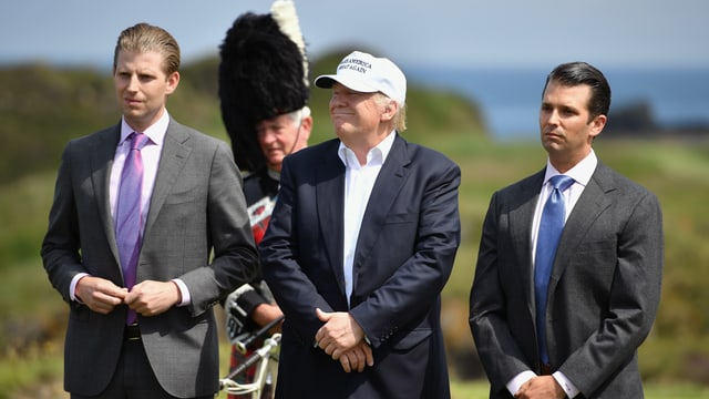 Trump mit weisser Schildmütze, seine Söhne stehen neben ihm. Dahinter ein Dudelsack-Spieler in Schotten-Uniform. Trump mit weisser Schildmütze, seine Söhne stehen neben ihm. Dahinter ein Dudelsack-Spieler in Schotten-Uniform.