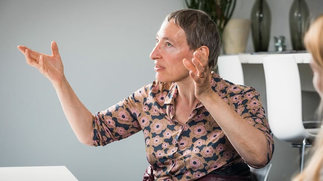 Eine Frau mit kurzen grauen Haaren und Blumenbluse gestikuliert mit beiden Händen. Eine Frau mit kurzen grauen Haaren und Blumenbluse gestikuliert mit beiden Händen.