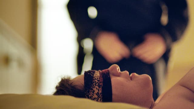 Frau mit verbundenen Augen auf Bett, man sieht nur ihr Gesicht, im Hintergrund öffnet ein Mann seine Hose. Frau mit verbundenen Augen auf Bett, man sieht nur ihr Gesicht, im Hintergrund öffnet ein Mann seine Hose.