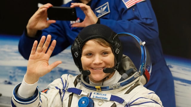 Anne McClain in Astronautenuniform, winkt ins Bild. Anne McClain in Astronautenuniform, winkt ins Bild.