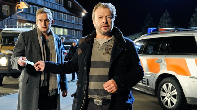 Zwei Männer stehen vor Polizeiwagen. Der vordere reicht dem hinteren ein Stück Papier. Zwei Männer stehen vor Polizeiwagen. Der vordere reicht dem hinteren ein Stück Papier.