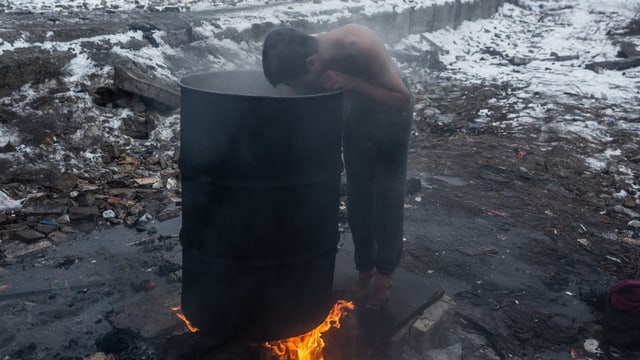 Ein Junge beugt sich in eine Abfalltonne, unter der ein Feuer brennt. Er trägt nur eine Hose und es liegt Schnee. Ein Junge beugt sich in eine Abfalltonne, unter der ein Feuer brennt. Er trägt nur eine Hose und es liegt Schnee.