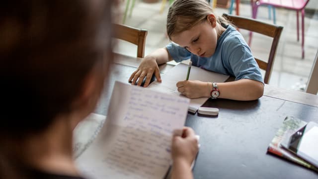Mädchen schreibt am Küchentisch, Mutter hält Papier in der Hand Mädchen schreibt am Küchentisch, Mutter hält Papier in der Hand