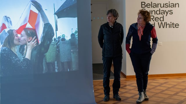 Blick in den Ausstellungsraum: Zwei Frauen schauen auf eine Leinwand, auf die eine Fotografie projiziert wird. Blick in den Ausstellungsraum: Zwei Frauen schauen auf eine Leinwand, auf die eine Fotografie projiziert wird.