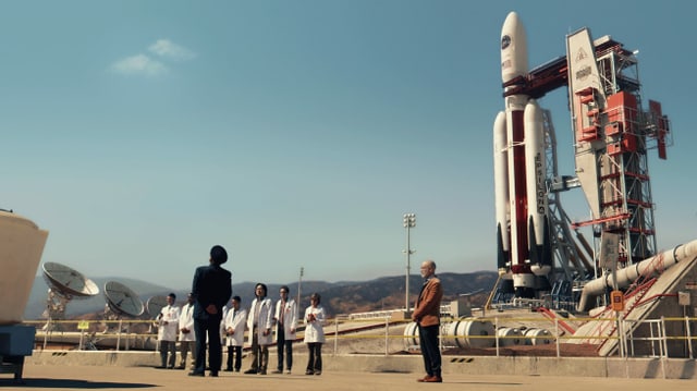 Von Weitem ein Platz, darauf steht eine Rakete und davor eine Gruppe winzig scheinender Menschen. Einer trägt eine Uniform, einer einen Anzug, die restlichen tragen weisse Kittel. Von Weitem ein Platz, darauf steht eine Rakete und davor eine Gruppe winzig scheinender Menschen. Einer trägt eine Uniform, einer einen Anzug, die restlichen tragen weisse Kittel.
