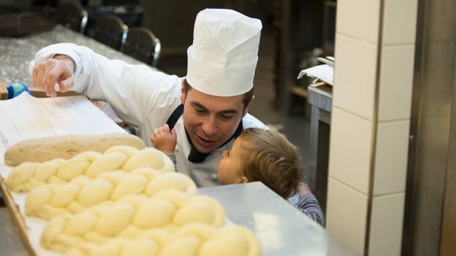 Ein Mann mit Kochhut ist dabei, Zöpfe zu backen und sein kleiner Sohn schaut zu. Ein Mann mit Kochhut ist dabei, Zöpfe zu backen und sein kleiner Sohn schaut zu.