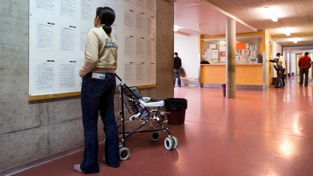 Frau mit Kinderwagen blickt an eine Wand mit einer Informationstafel. Frau mit Kinderwagen blickt an eine Wand mit einer Informationstafel.