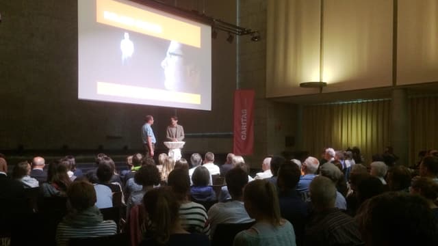 Saal voller Leute mit Podiumsdiskussion Saal voller Leute mit Podiumsdiskussion