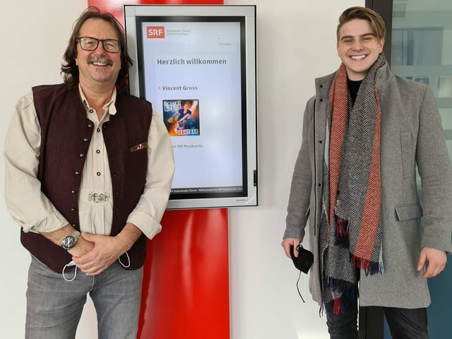 Vincent Gross steht neben Musikredaktor. Auf einem Display im Hintergrund ist ein Bild des Sängers mit der Überschrift «Willkommen» zu sehen. Vincent Gross steht neben Musikredaktor. Auf einem Display im Hintergrund ist ein Bild des Sängers mit der Überschrift «Willkommen» zu sehen.