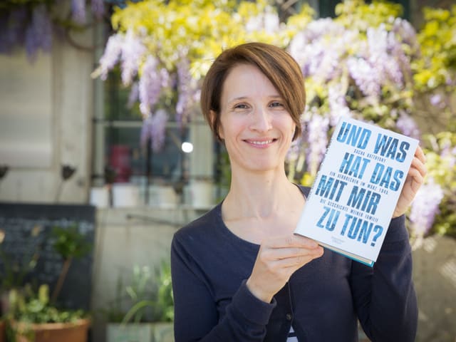 Eine Frau steht vor einem Fliederbaum und hält lachend ein Buch.