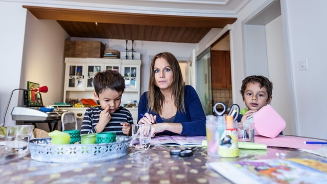 Eine Mutter sitzt am Wohnzimmertisch mit zwei Kindern. Eine Mutter sitzt am Wohnzimmertisch mit zwei Kindern.