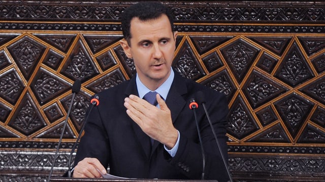 Assad hält eine Rede an einem Stehpult mit zwei kleinen Mikrofonen. Assad hält eine Rede an einem Stehpult mit zwei kleinen Mikrofonen.