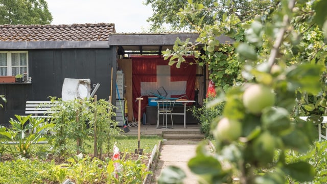 Ein Schrebergartenhäuschen mit geschützem Sitzplatz, an dessen Rückwand eine grosse Schweizerfahne hängt. Ein Schrebergartenhäuschen mit geschützem Sitzplatz, an dessen Rückwand eine grosse Schweizerfahne hängt.