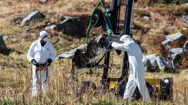 Zwei Unfallermittler in weissen Anzügen bergen einen Teil des Helikopterfahrgestells, das an einem Kran hängt Zwei Unfallermittler in weissen Anzügen bergen einen Teil des Helikopterfahrgestells, das an einem Kran hängt