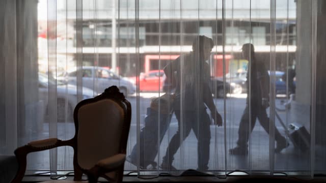 Blick aus einer Hotellobby durch ein Fenster mit Vorhang auf zwei Touristen, die auf dem Trottoir ihre Rollkoffer ziehen. Blick aus einer Hotellobby durch ein Fenster mit Vorhang auf zwei Touristen, die auf dem Trottoir ihre Rollkoffer ziehen.