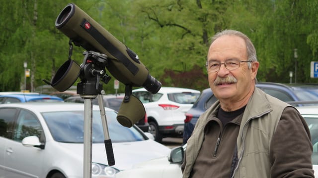 Ein Mann steht auf einem Parkplatz, neben einem grossen Fernrohr auf einem Stativ. Ein Mann steht auf einem Parkplatz, neben einem grossen Fernrohr auf einem Stativ.