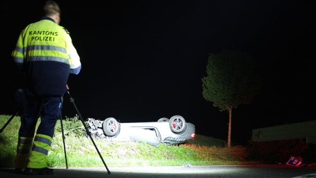 Auto liegt auf Dach neben Strasse, im Vordergrund Polizist mit Kamera Auto liegt auf Dach neben Strasse, im Vordergrund Polizist mit Kamera